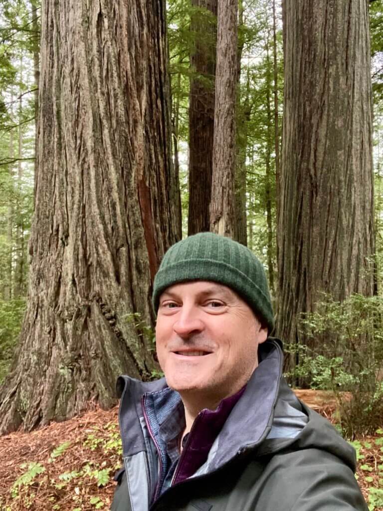 Matthew Kessi poses for a selfie in front of several large coast redwood trees.  He's smiling and wearing a green beanie and green raincoat.  Exploring a redwood forest is a great thing to do in Brookings Oregon.  