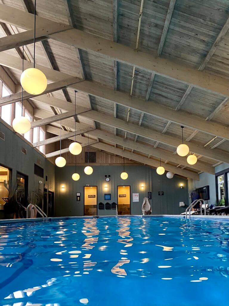 An indoor swimming pool glows with bright blue water while the round lights cast a yellowish glow on the surface of the water. High wood beams lead up to windows shining in with bright light.