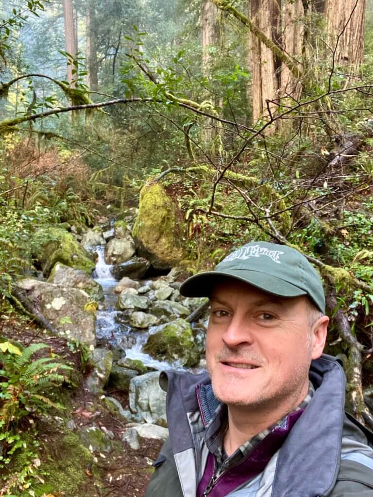 Matthew Kessi poses for a selfie on a hike through Redwoods in Oregon.  Behind him is a babbling creek navigating through medium sized boulders and redwoods and fir trees rising up in the distance. 