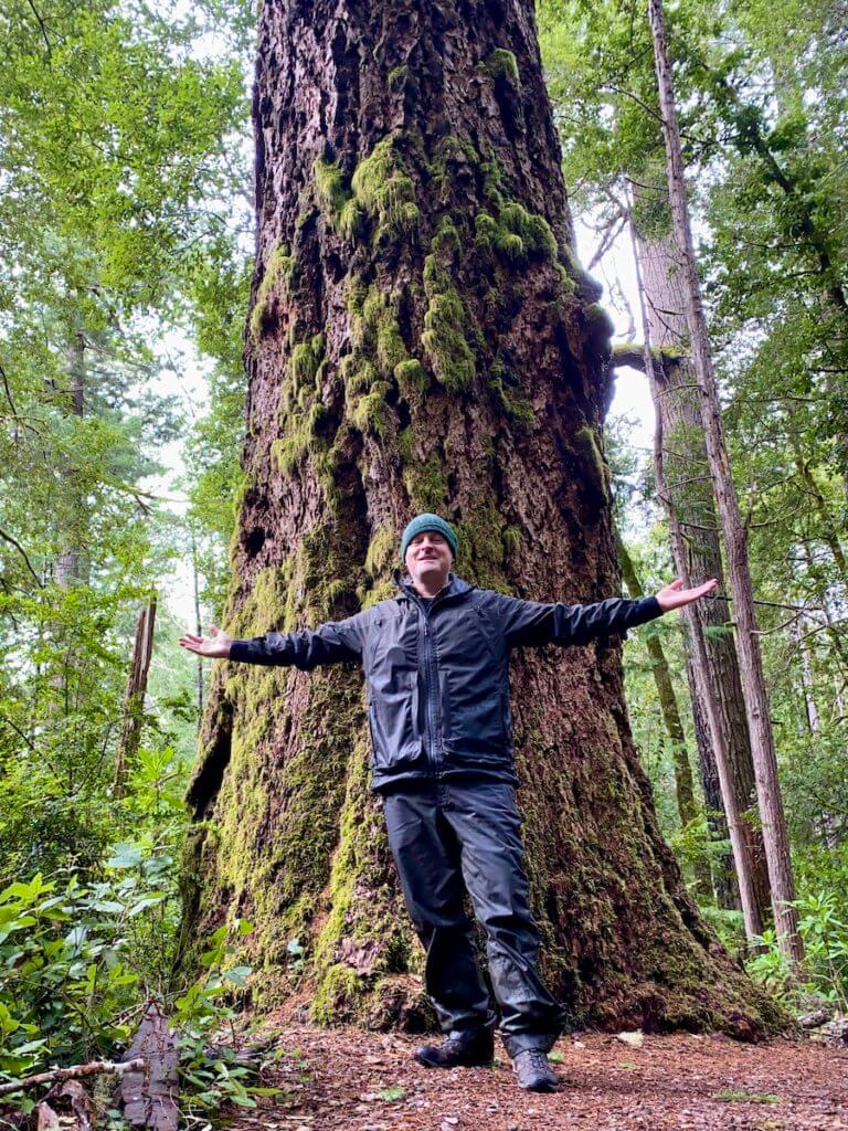 Matthew Kessi stands, arms stretched out under a giant Douglas fir tree near Gold Beach on the Oregon Coast. He's wearing rain gear and a green beanie and smiling at the grandeur of the dense green forest.