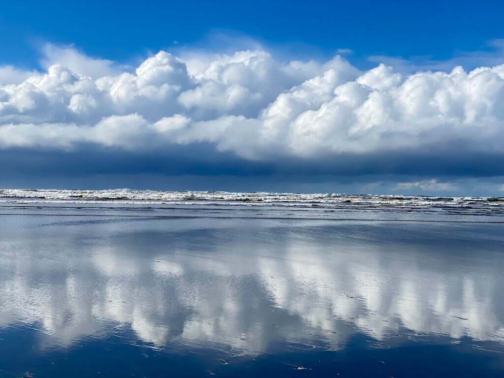 Brilliant blue sky reflects off a glistening sandy beach near an Oregon Coast town. The clouds are puffy white while the surf is crashing on the beach.