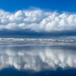 Brilliant blue sky reflects off a glistening sandy beach near an Oregon Coast town. The clouds are puffy white while the surf is crashing on the beach.