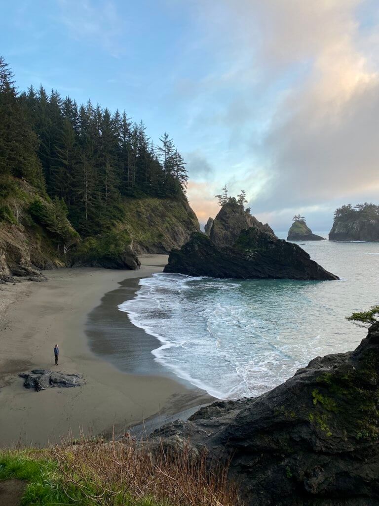 Secret Beach along the Southern Oregon Coast is mystical in the waning sunset glow. The sky is a mixture of yellow and orange and purple and a man stands on the smooth sandy beach looking at the light waves foaming as they hit the shore.