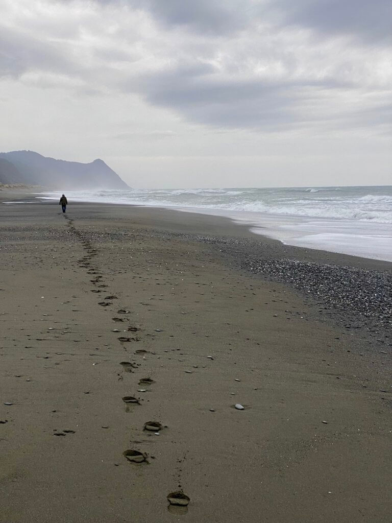 The footprints of a beach walker flow off into the horizon with a small person walking near the surf of the Pacific Ocean along the Oregon Coast.  The gray clouds in the sky swirl around. 