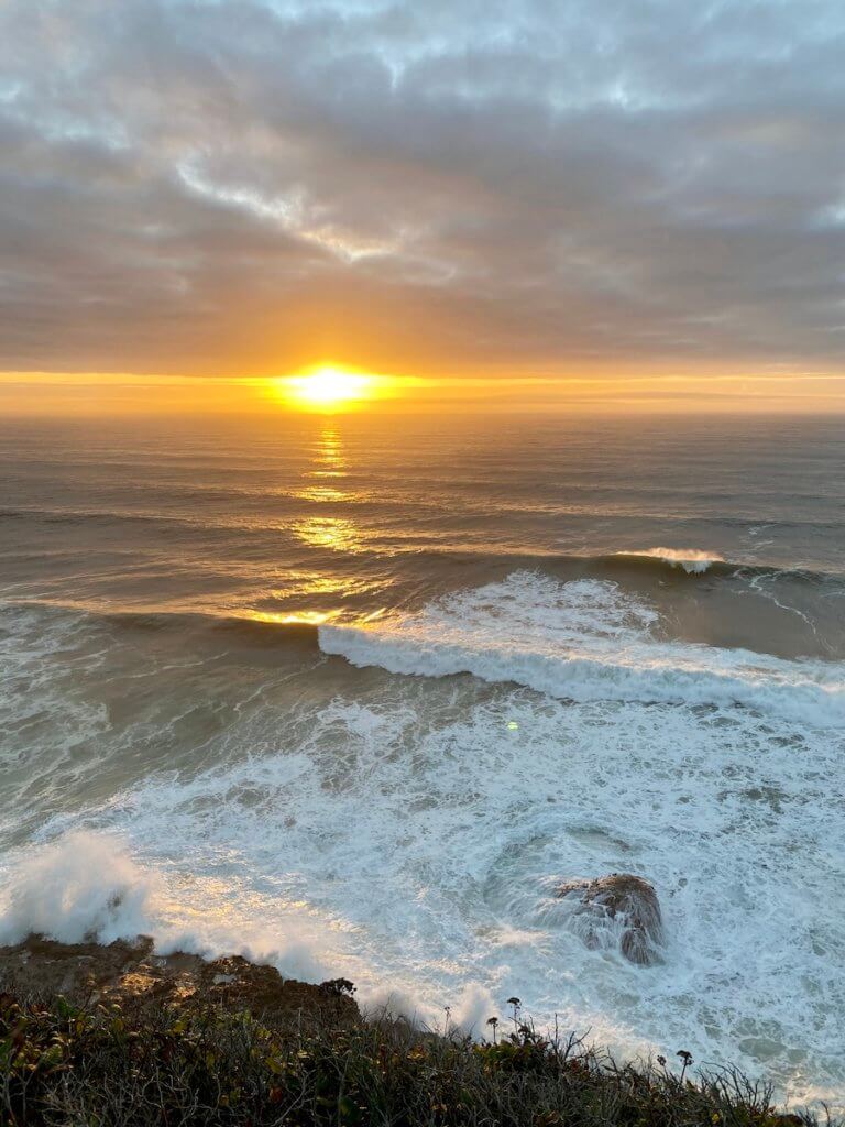 A buttery glow of sun begins to fade over the horizon of the Pacific Ocean as foamy white waves crash against rocks.