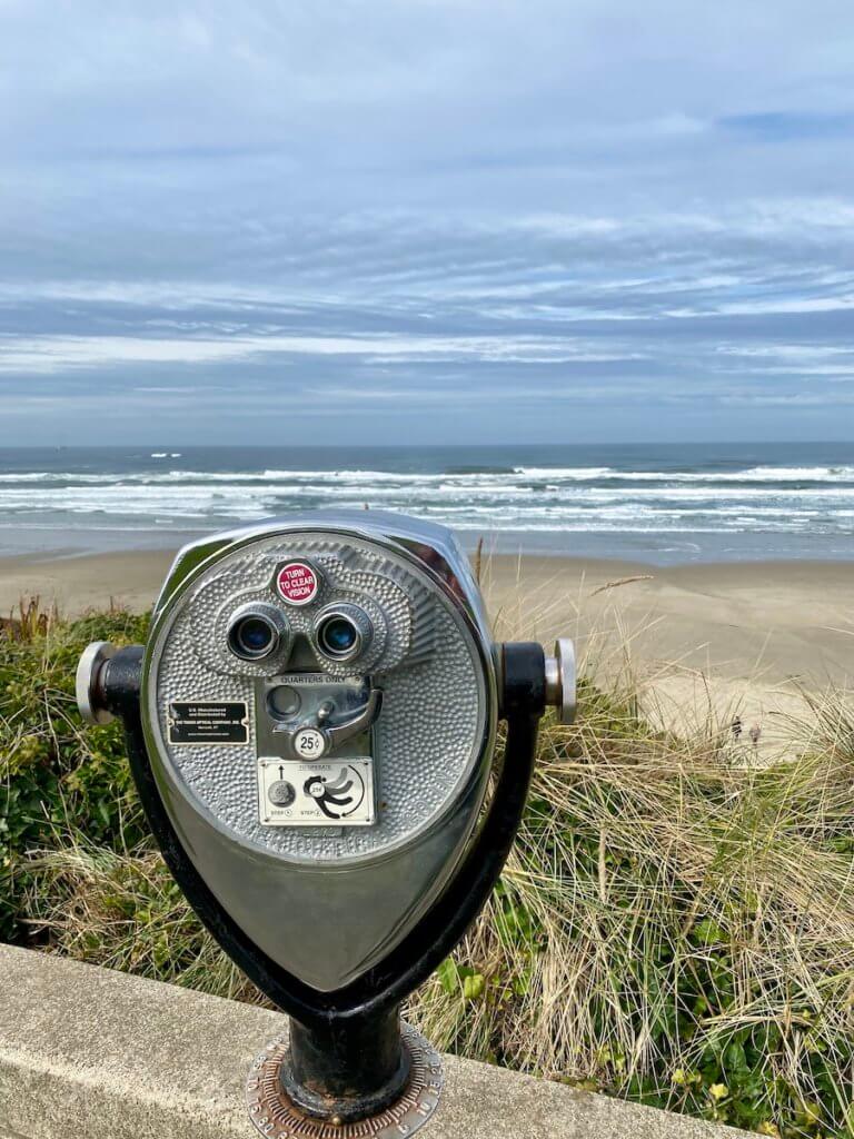A rustic stainless steel fixed binocular looks out at the surf in Newport, which is one of the best Oregon Coast Towns for beaches. The white caps of the waves in the Pacific Ocean roll into the sand while the gray clouds seem to swirl above.