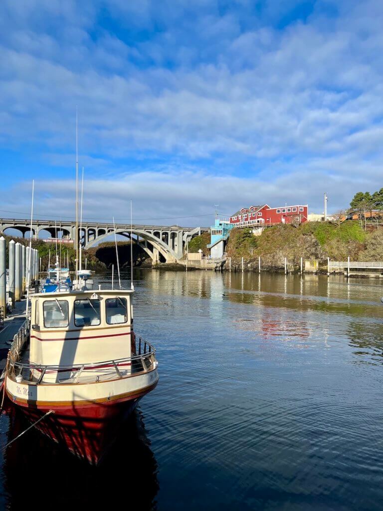 Depoe Bay is one of the best Oregon Coast Towns for day trips from the Willamette Valley. Here, a red hulled boat sits in the harbor where the iconic Art Deco bridge spans in the background and a rustic red building with white trim sits atop the harbor. The sky is blue with white swirls.