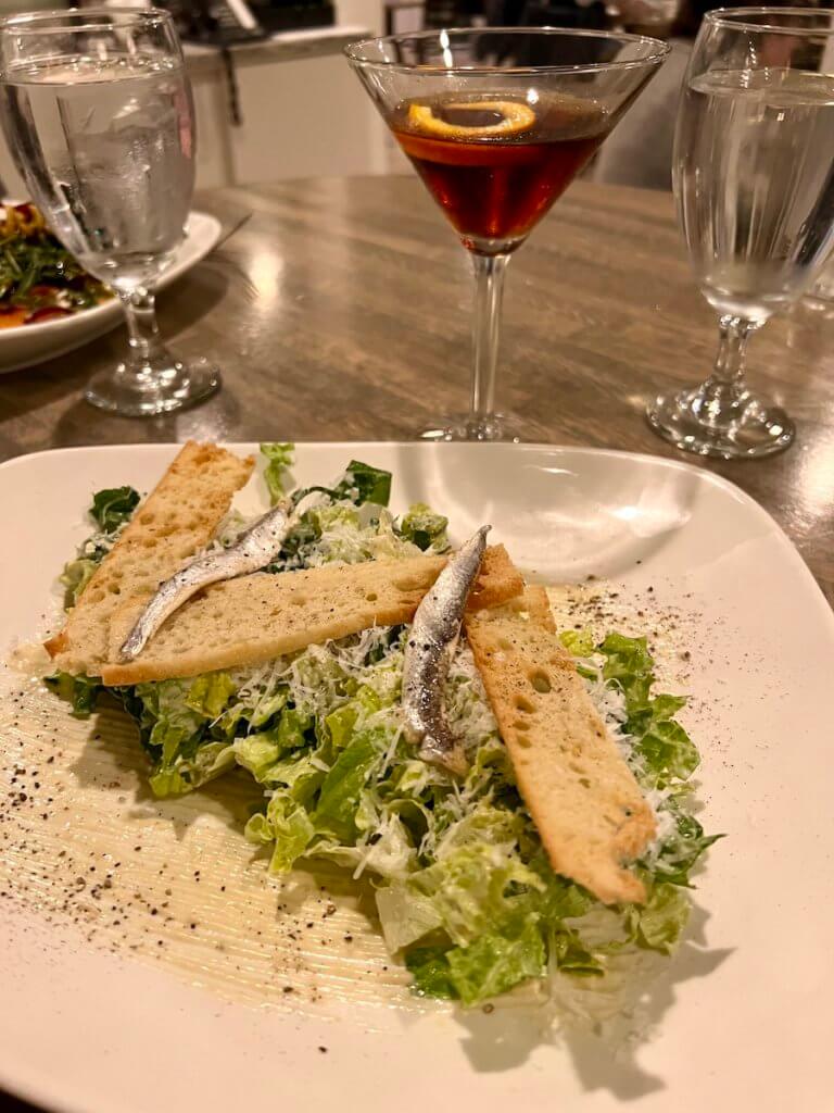 A Manhattan is half consumed next to a plate of Caesar salad with whole anchovy fish atop. This appears to be at a fine dining establishment.
