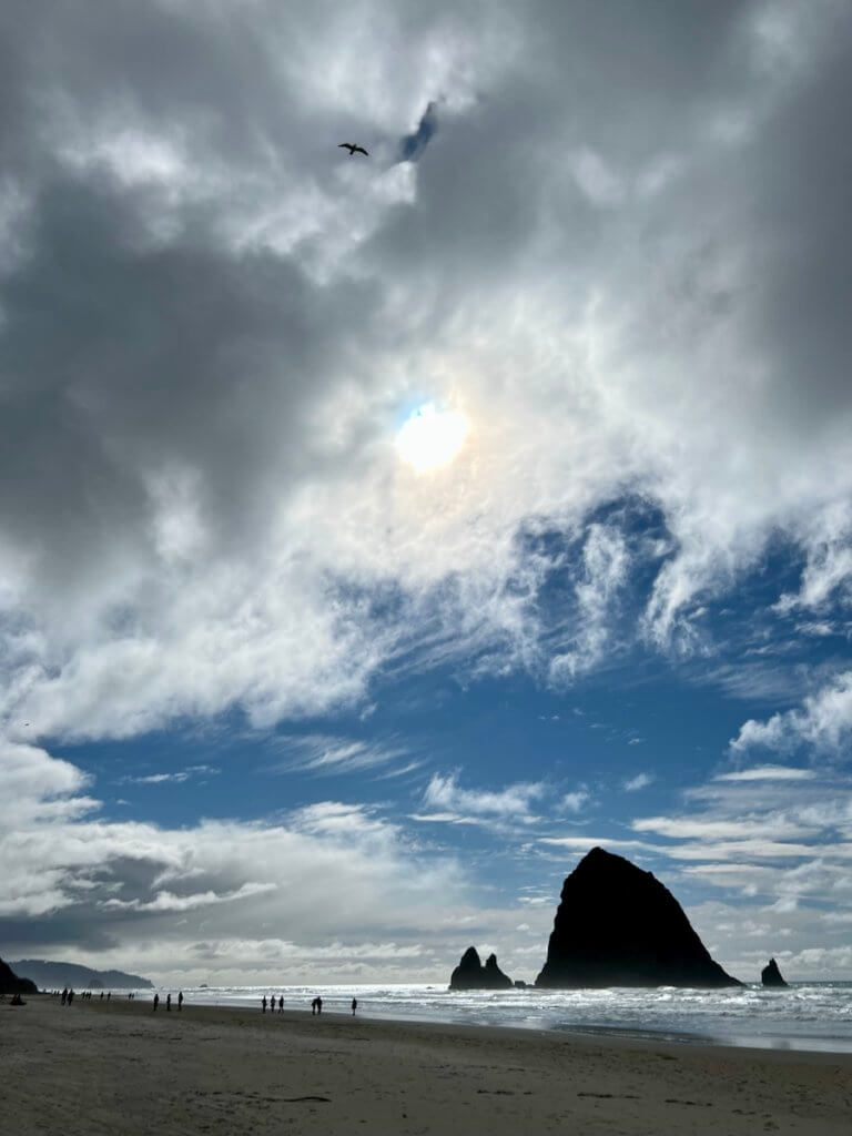 People walk on the beach toward iconic Haystack Rock, in Cannon Beach, Oregon. The sky is blue with some clouds and the sun pokes through to shine on an open sandy beach.