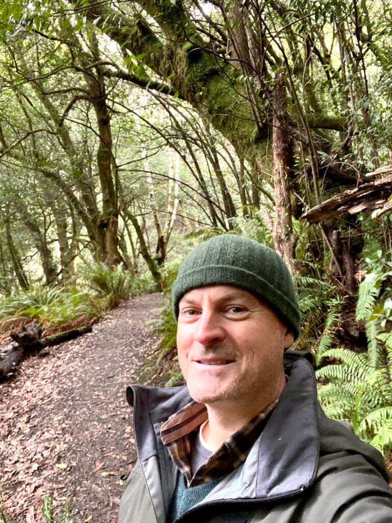 Matthew Kessi smiles in the middle of a Myrtlewood grove near one of the best Oregon Coast Towns of Gold Beach. He's wearing a green beanie and smiling.