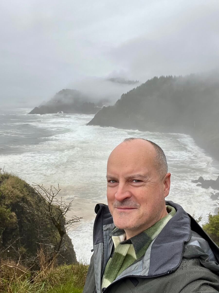 Matthew Kessi poses for a selfie overlooking the stormy surf far below a cliff near the Oregon Coast town of Florence. There is mist drafting up from the hill of fir trees.