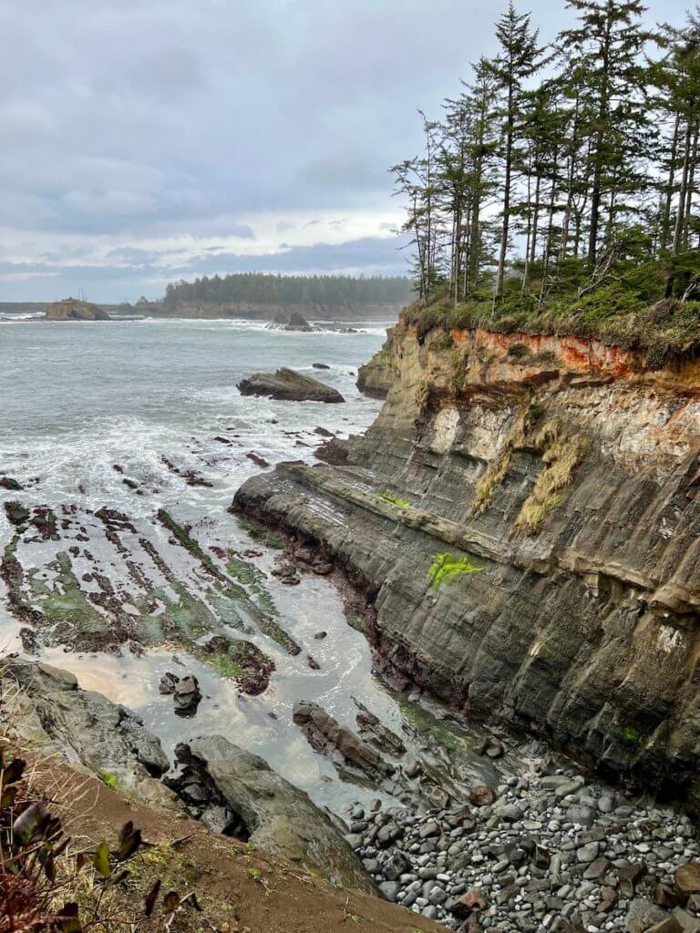 A seascape along the Oregon Coast reveals rocks worn away by years of waves crashing. Atop the cliff are fir trees clinging to orangish rocky soil and the skies are gray.