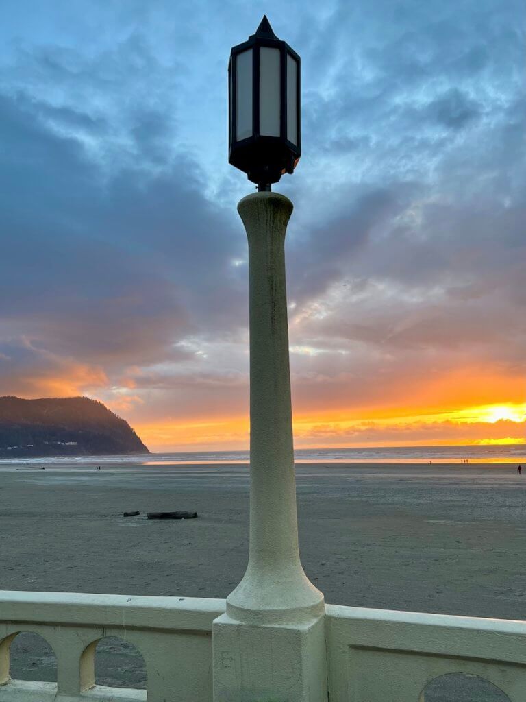 Seaside is one of the best Oregon Coast Towns for a wide open beach. Here the sunset is glowing orange and yellow while people walk on the sand in the far distance. A lamppost on the Seaside Promenade is not yet lit for the evening.