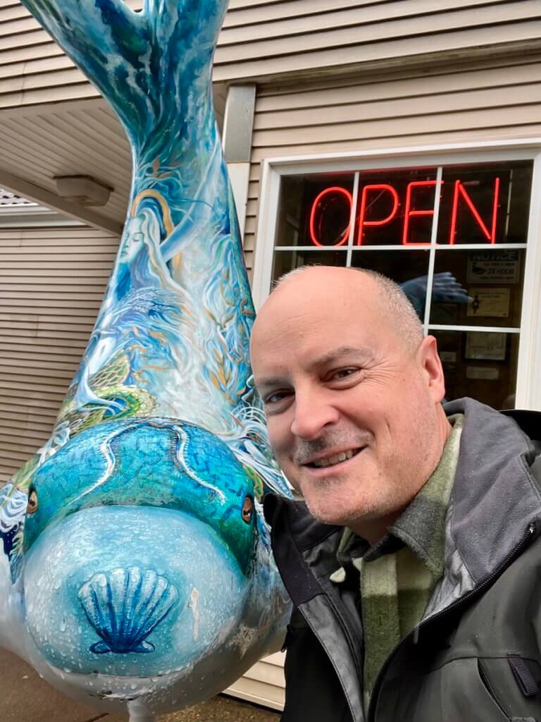 Matthew Kessi poses for a selfie with a painted blue sealion cub. Behind them is a neon Open sign. he's smiling and wearing a green plaid shirt.