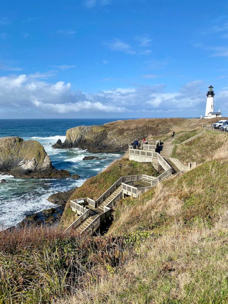 Yaquina Head lighthouse is in the Oregon Coast town of Newport. Stairs lead down to the rocky beach with waves crashing from the sea. In the distance the lighthouse rises up into a blue sky.