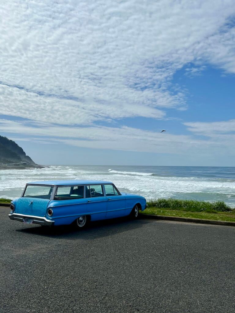 A vintage blue station wagon parks at the edge of a cliff overlooking the waves of the Pacific Ocean. There is a seagull flying through the sky which is blue with swirls of clouds.