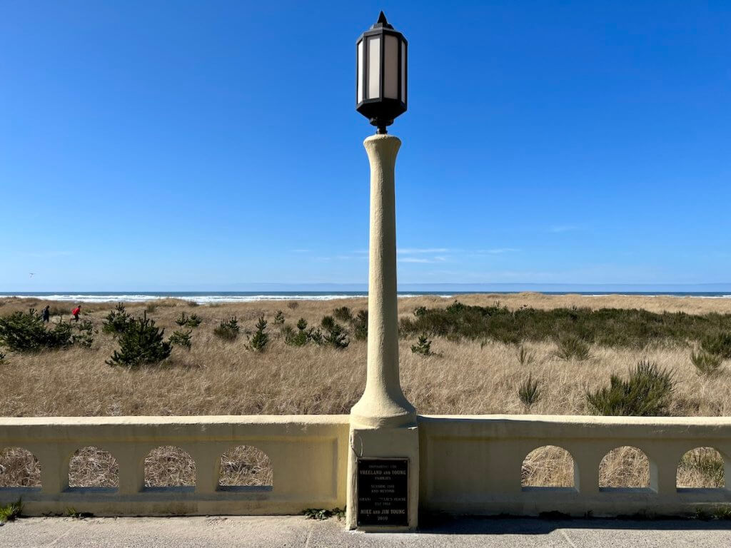 The iconic lamp post on the Seaside Promenade is made of yellow painted concrete with a black casing for the light fixture. The sky is bright blue and just a faint line of surf from the ocean can be seen banging onto the beach.