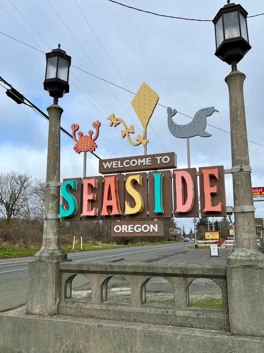 The welcome sign to Seaside Oregon is festive with different colored beach style letters perched between two historic looking lamp posts. There is a orange crab, a yellow kite, and a gray seal above the sign.