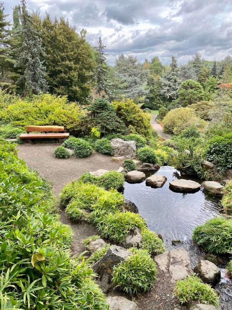 The layers of nature connect the viewer to water, rocks, trees, flowers and a bench made from wood. The sky is a dramatic mix of gray clouds and some patches of blue. A red moon-bridge can be seen far in the distance of this Japanese themed garden in Seattle.
