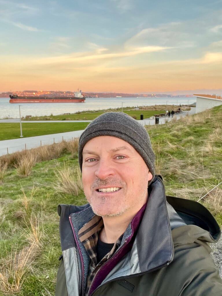 Matthew Kessi poses for a selfie at Dune Peninsula at Point Defiance Park. He's wearing a gray cap and smiling big while a ship is resting in the water behind him. He is showing participants how to nature forward while on vacation by leading them around the park.