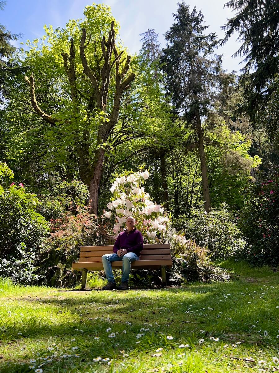 Matthew Kessi sits on a park bench in the Rhododendron Garden at Point defiance Park. There is a brightly colored flowing bush behind him and a variety of green fir and maple trees in the canopy above.