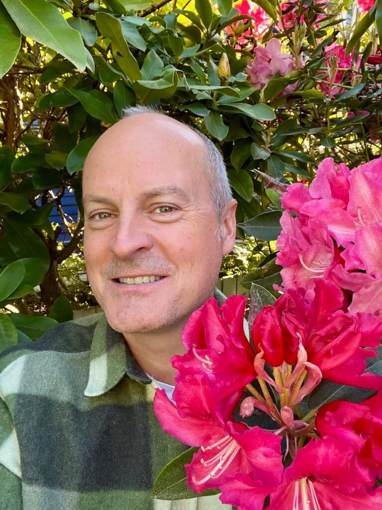 Matthew Kessi poses for a selfie in a large rhododendron bush with bright red and pink blooms. he's smiling and wearing a green and white checkered shirt.