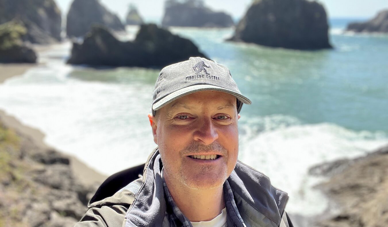 Matthew Kessi poses above a sandy beach on the OREGON COAST with waves crashing around large rock stacks in the background. He's wearing a grey cap and smiling big.