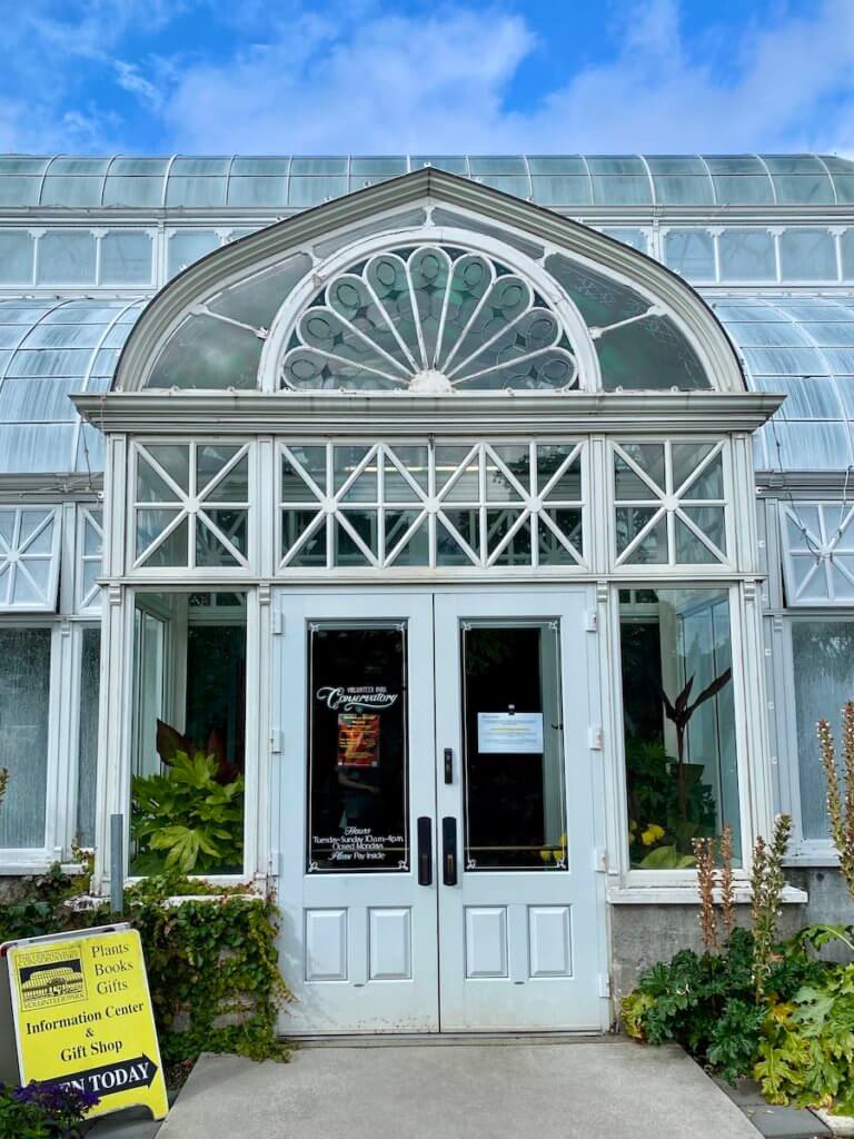 The Volunteer Park Conservatory is iconic with the metal and glass Victorian look under a blue sky day. The doors leading into the building are white and the sandwich board sign is yellow with black lettering.