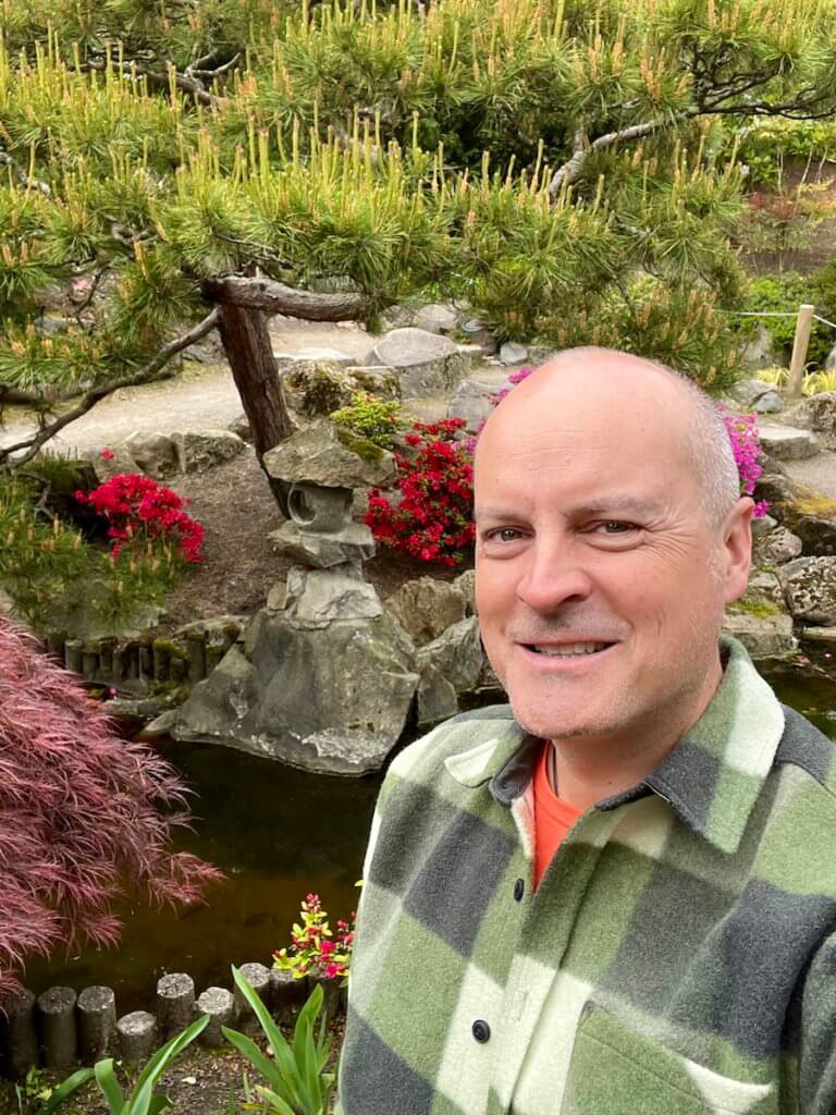 Matthew Kessi poses for a selfie in front of a Japanese style lantern in the middle of a garden in Seattle. He's smiling and wearing an orange t-shirt and green checkered shirt. There is a red lace leaf maple as well as bright red azaleas blooming in the background.