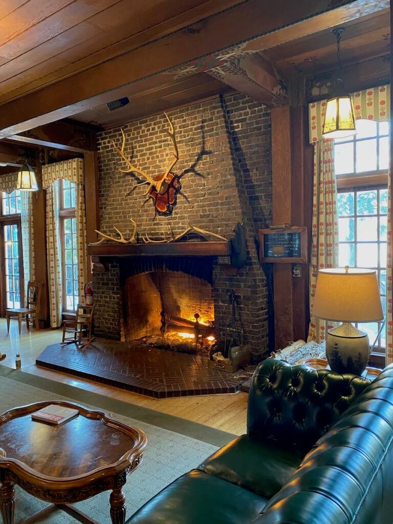 The inside of Lake Quinault Lodge shows a roaring fire and a cozy green leather couches while a rack of antlers from a Roosevelt Elk adorn the mantle.