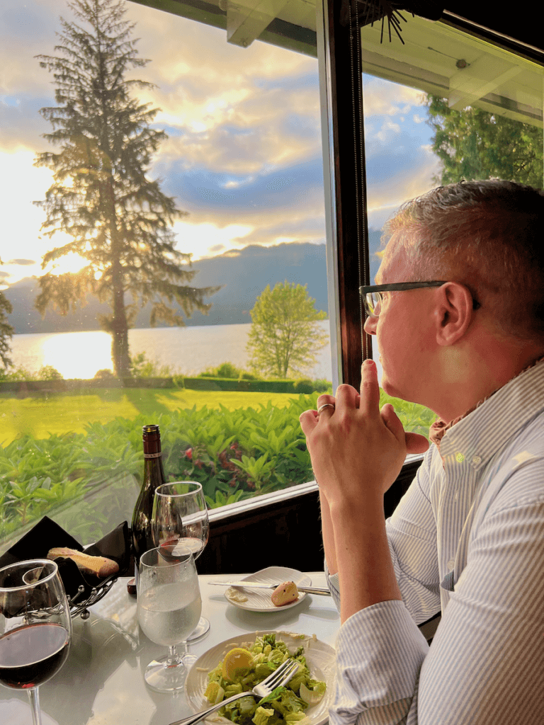 A man looks out from his table to Lake Quinault while the sun sets over the peaceful lake casts a buttery glow. He is wearing glasses and a striped button down shirt. There are two glasses of red wine on the table and a Caesar salad on the table with a silver fork.