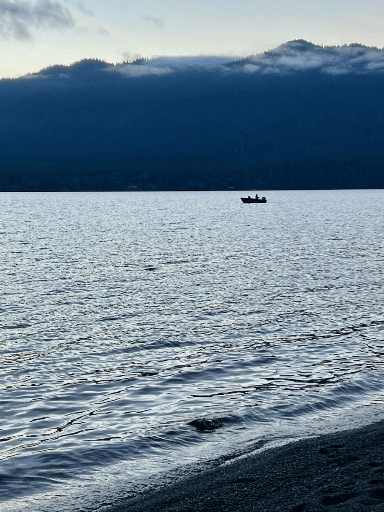 A fishing boat is quietly lurking on the horizon of Lake Quinault while small waves wash onto the shore. The mountains in the distance have mist under a blue sky.