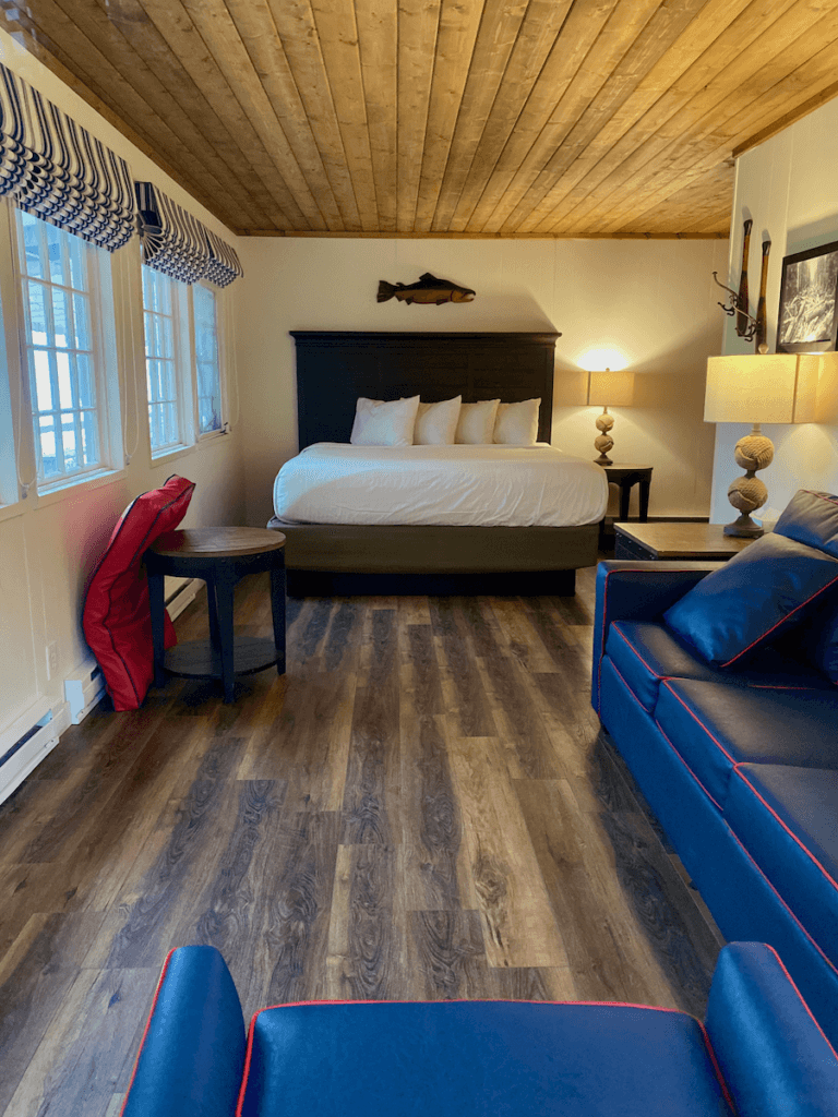 A boat house room at Lake Quinault Lodge shows fresh appointments including a plush queen sized bed and blue couch and chair with red stripe trim.