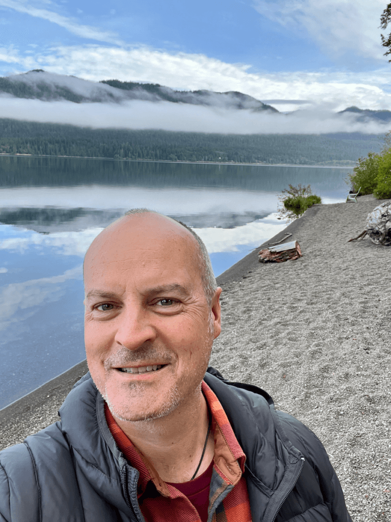 Matthew Kessi poses for a selfie on the bank of Lake Quinault. The beach is made of pebbles and the water is flat and reflects the outlines of the mountains in the background under blue sky.
