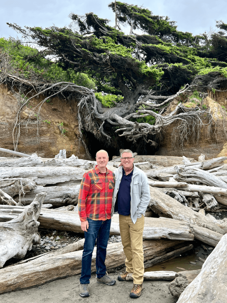 Matthew Kessi and Joe pose for a shot in front of the Tree of Life near Kalaloch Lodge in the Olympic National Park. Matthew is wearing an orange check shirt and blue jeans while Joe is wearing tan hiking pants and a gray jacket.