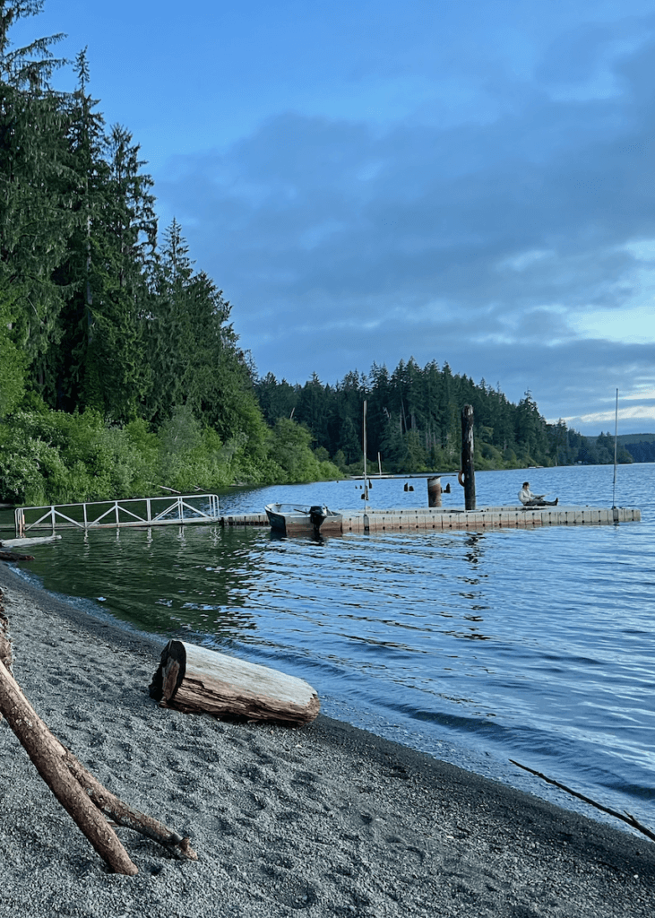 The quiet waters of Lake Quinault lap onto a pebble rock beach while a man sits on a dock in the distance with a fishing pole. The sky is gray blue and there is a large piece of log on the beach.