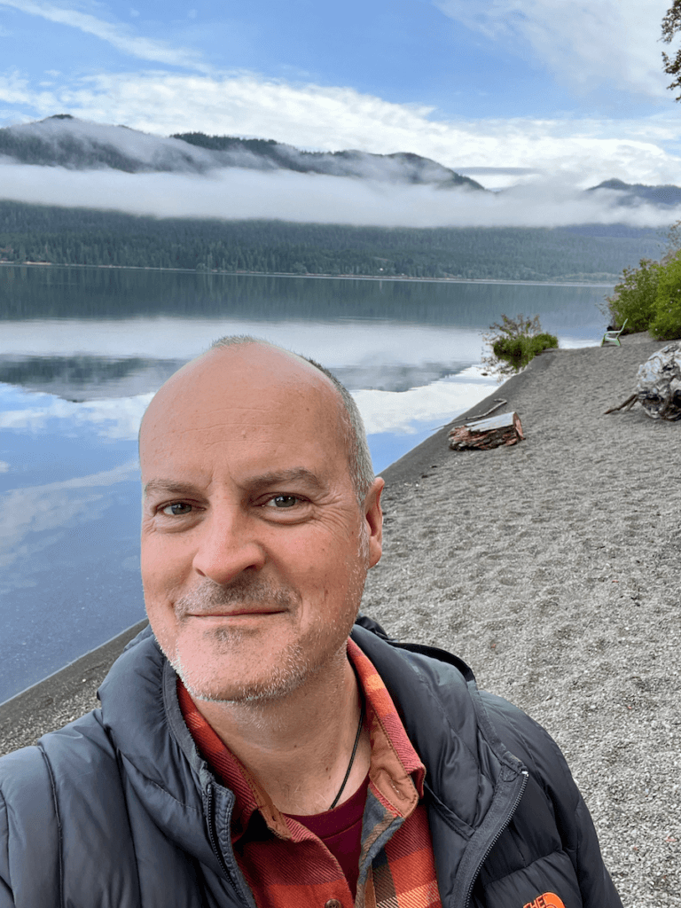 Matthew Kessi takes a selfie in from of a quiet Lake Quinault while low clouds float through the hills in the background under blue sky.