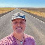 Selfie of Matthew Kessi on an open road in Eastern Oregon. He's smiling and wearing a gray cap and red shirt with a necklace holding a round mirror. There is a wheat field on one side of the road and a plowed dirt field on the other. In the distance is a wind farm.