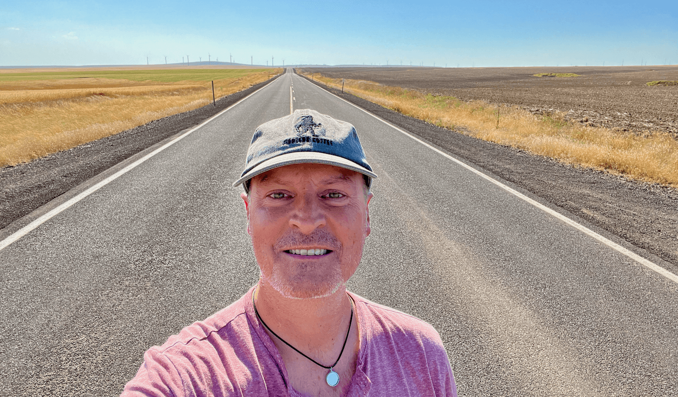 Selfie of Matthew Kessi on an open road in Eastern Oregon. He's smiling and wearing a gray cap and red shirt with a necklace holding a round mirror. There is a wheat field on one side of the road and a plowed dirt field on the other. In the distance is a wind farm.