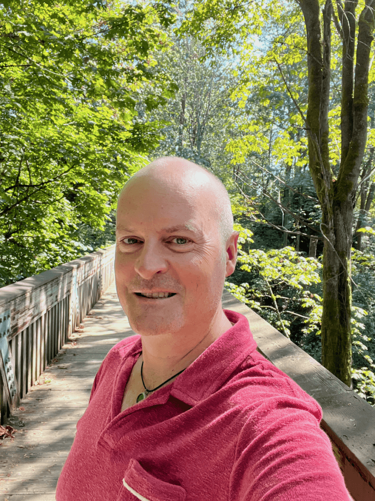 Matthew Kessi poses for a shot in Ravenna Park. He's leading people on a curated nature connection over a footbridge. He's wearing a coral red shirt and smiling while the maple tree leaves float in the blue sky above.