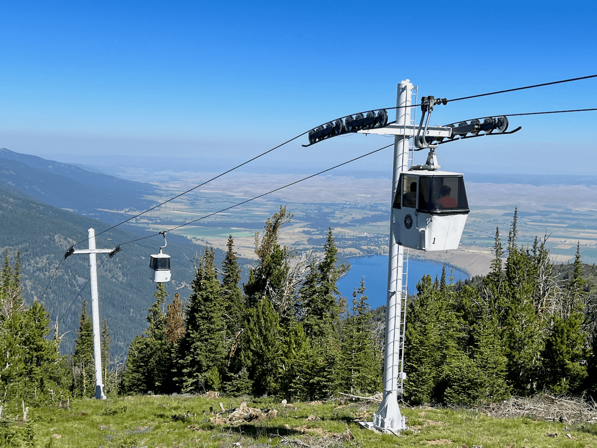 View from the summit of Wallowa Lake Tramway. The blue lake is in the background with the rolling farmland of Joseph in the background. There are two white gondolas gliding on black cables.