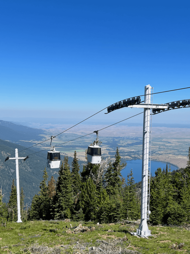 View from the summit of Wallowa Lake Tramway. The blue lake is in the background with the rolling farmland of Joseph in the background. There are two white gondolas gliding on black cables.