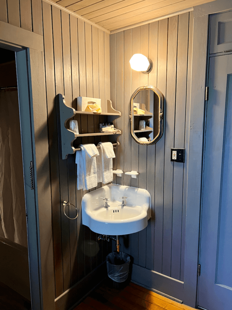 Inside a room at the Wallowa Lake Loge, the corner is painted blue on wood boards and there is a corner sink, complete with towels and soap and a mirror affixed to the wall. A glowing light is turned on above the scene.