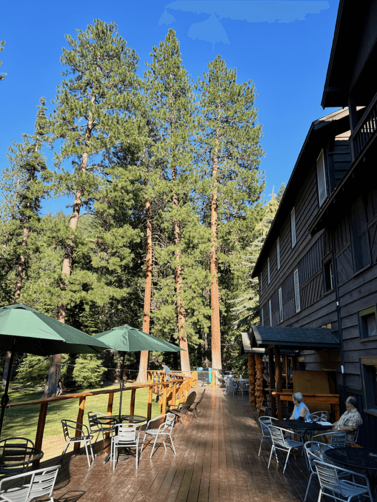 The porch of the Wallowa Lake Lodge has a comfortable array of tables and chairs, some are covered by umbrellas. In the distance several ponderosa pine trees rise up to the blue sky above.
