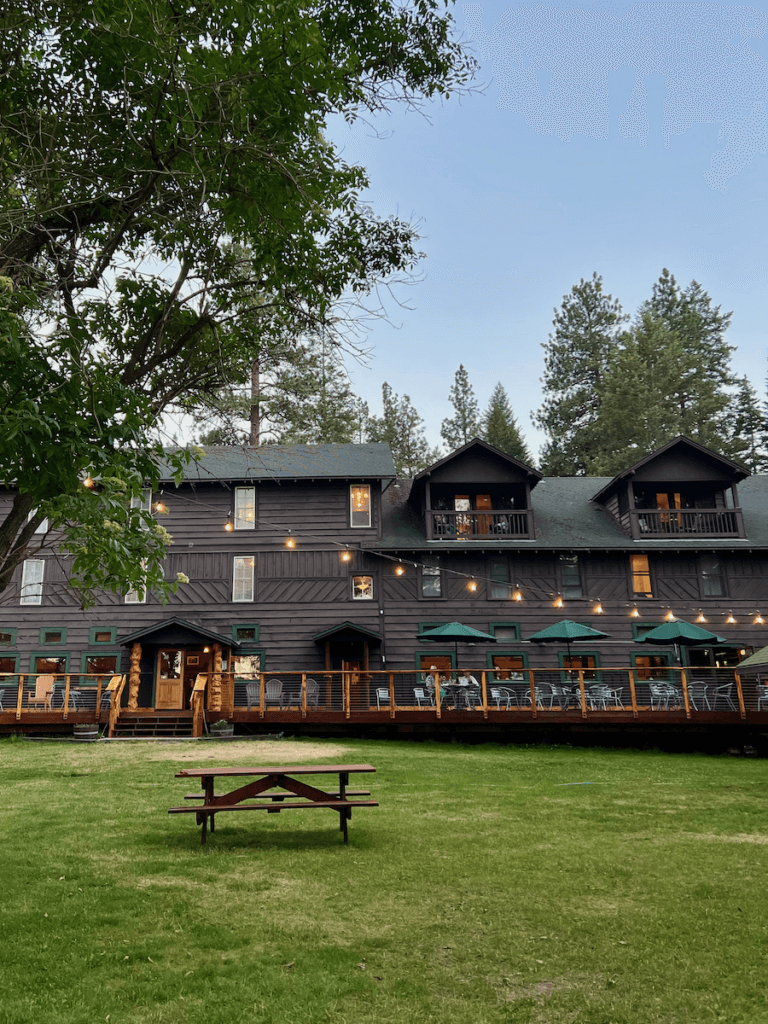 Wallowa Lake Lodge is a comfortable place and starts to glow around sundown. Here a string of lights shines over a deck area with tables and umbrellas where people are enjoying a beverage and the view of the lawn. There is a lone picnic table in the foreground on the green grass.