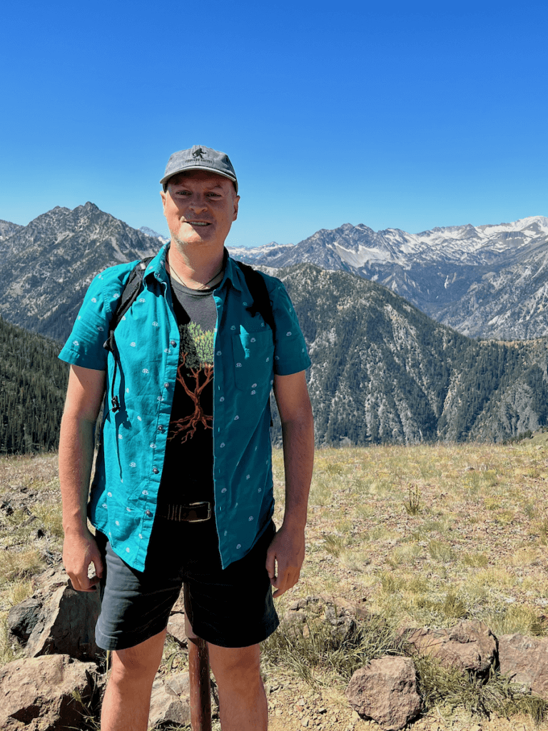 Matthew Kessi stands atop Howard Mountain, steps from the Wallowa Lake Tramway. Impressive mountain peaks rise up in the distance, jagged with rock and some snow. he's smiling and wearing a gray cap and a blue button down shirt with navy shorts.