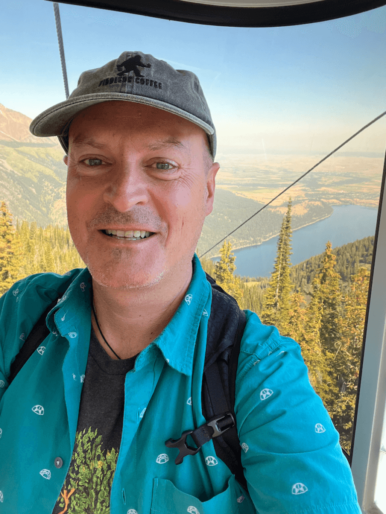 Matthew Kessi grins as he rides a gondola to the top of Howard Mountain. In the background Wallowa Lake shines blue and cables from the tram cane be seen. Matthew is smiling brightly and wearing a gray cap.