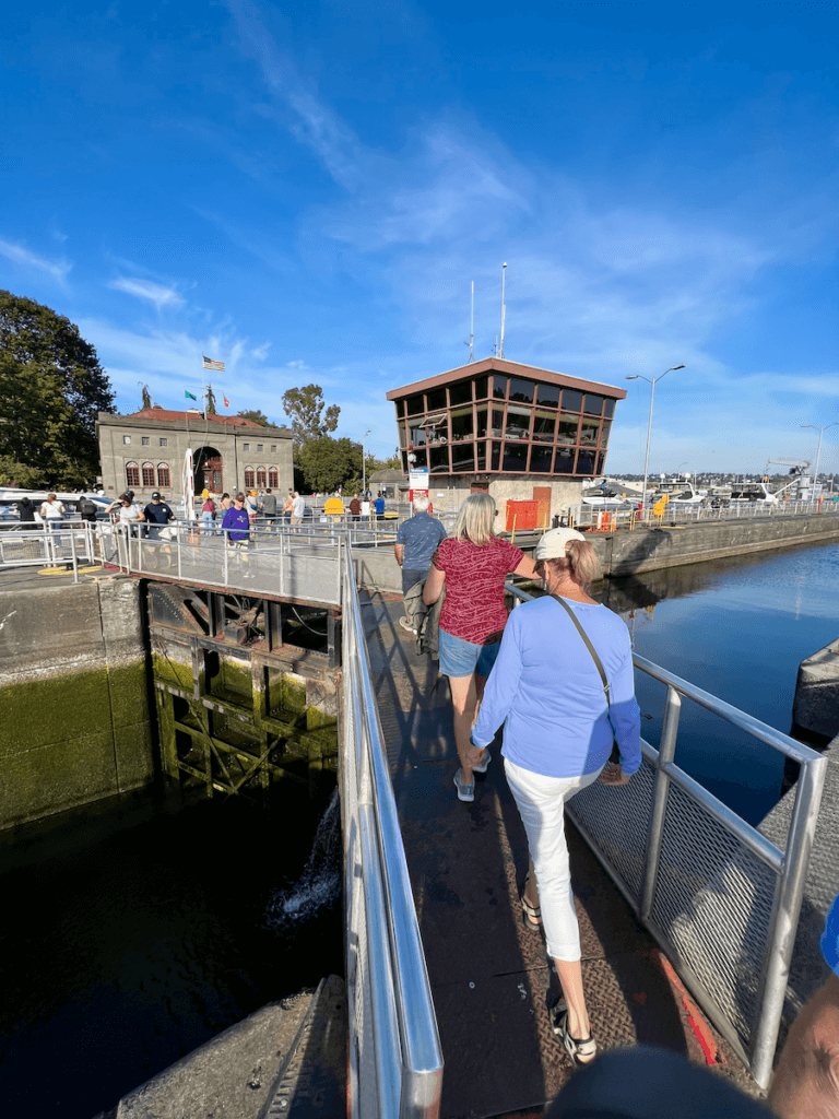 Tourists walk across a huge gate separating freshwater from saltwater at Ballard Locks. There are three people crossing and in the background you can see the Army corps of Engineers buildings.