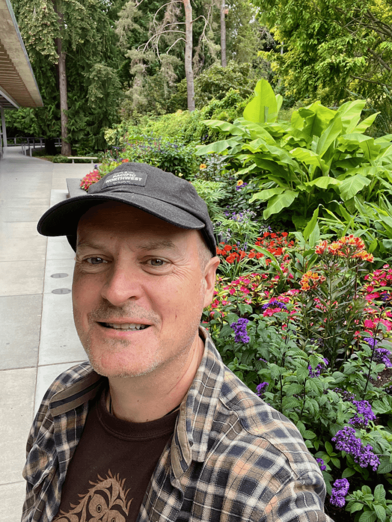Matthew Kessi poses for a selfie at the entrance to Bellevue Botanical Garden. He's wearing a black hat and a plaid brown shirt and smiling in front of a flower planter full of different colored flowers and various textures of green plants.