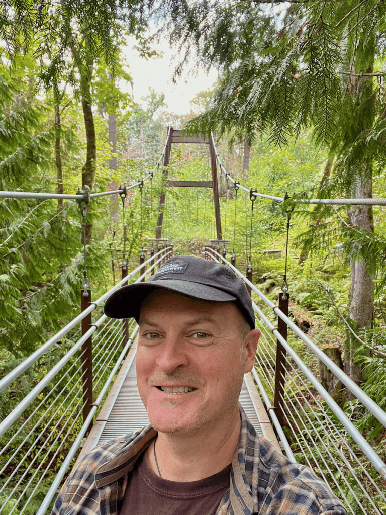 Matthew Kessi poses for a selfie on the suspension bridge at the Ravine Experience in Bellevue Botanical Garden. He's smiling big and wearing a black cap and brown plaid shirt. The green trees surround him and you can see the thick cables of the bridge behind him.